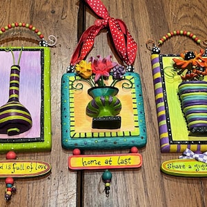 May include: Three colorful wooden signs with painted flowers and vases, each with a different message: "my world is full of love", "home at last", and "share a giggle today".