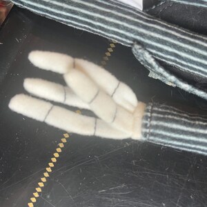 以下が含まれることがあります： 黒と白のピンストライプ柄の布製袖が付いた、ぬいぐるみの腕と手。手はオフホワイトのフェルト素材で作られており、指と関節を表現する細い黒いステッチが施され、ストライプ柄の袖口から伸びています。