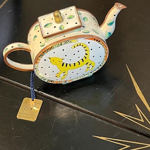 May include: A decorative metal teapot featuring a cream-colored body with green leaf patterns and a gold-toned lid knob. The side displays a yellow tiger illustration on a white background with black dots, framed by an orange and green border.