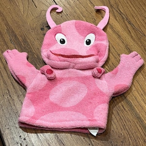 May include: A pink plush puppet with a smiling face, large white eyes, and two curved antennae. The puppet has a body with large pink polka dots and two arms with three fingers each. The puppet is made of a soft, textured material.