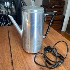 May include: A vintage, brushed aluminum coffee percolator with a black handle and a glass top. The electric coffee maker has a long spout and a black power cord. The percolator sits on a wooden surface.
