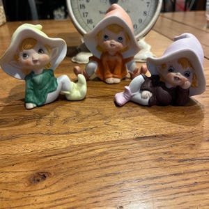 May include: Three ceramic figurines of seated elves with large hats. The elves have rosy cheeks, blonde hair, and are wearing different colored outfits: green, orange, and brown. The hats are in shades of white, peach, and lavender. A vintage scale is in the background.