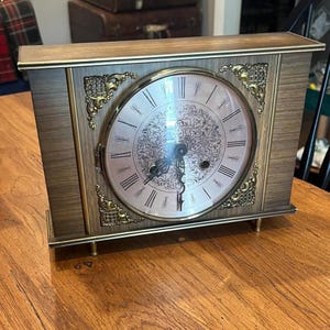 May include: A vintage wooden mantel clock with a light pink face and gold-colored accents. The clock features Roman numerals and ornate detailing around the face and on the sides. The clock is rectangular and sits on small brass feet.