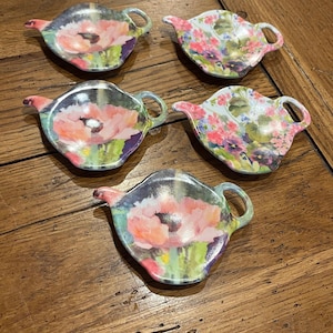 May include: Five decorative ceramic teapot-shaped coasters. Each coaster features a floral design with pink, red, and purple flowers. The coasters have a handle and spout, and are set on a wooden surface.