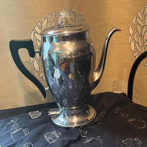 May include: A polished silver-tone metal electric percolator coffee pot featuring a tall, curved spout, a black handle, and a clear glass knob on the lid. The base includes a small power connector port, and the body has a reflective, mirror-like finish.