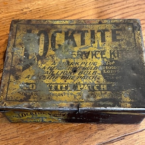May include: A vintage Locktite Service Kit tin, rectangular with a yellow and black color scheme. The tin's surface shows wear and tear, with text including "Locktite" and "Service Kit". The kit includes a spark plug, headlight bulb, and tire patch.