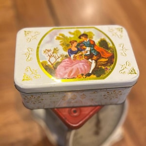 May include: A rectangular white tin with a hinged lid. The lid features a central oval image of a couple in period clothing, surrounded by a gold border. The tin is decorated with gold floral and scroll designs.