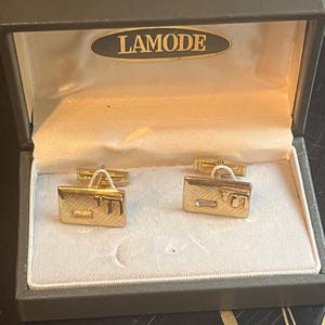 May include: A pair of gold-tone rectangular cufflinks featuring a textured surface, a small clear stone, and the Hebrew letters Chai. The cufflinks are presented in a grey hinged box with the brand name LAMODE printed on the interior lid.