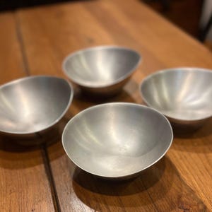 May include: Four small, oval-shaped metal bowls with a brushed silver finish are arranged on a wooden surface. The bowls have a simple, minimalist design, and are likely used for serving or display.