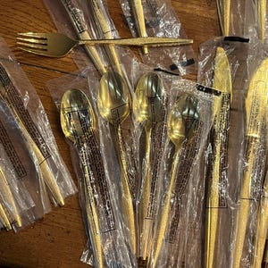 May include: A collection of gold-colored flatware, including forks, spoons, and knives, packaged in clear plastic. The utensils have a textured finish and are arranged on a wooden surface.