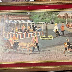 May include: A framed painting depicting a bustling city scene with a cafe called "Jon Maisie Cafe". The painting features a horse-drawn carriage, people walking, a car, and a bicycle. The painting is set against a backdrop of buildings and trees.