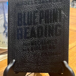 May include: A vintage book titled "Blueprint Reading for the Building Trades" is displayed on a black stand. The book has a textured, dark cover with the title in bold, dark lettering. The book is likely a manual or guide for construction.