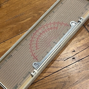 May include: A vintage drafting tool with a clear plastic ruler and a red protractor. The tool has a metal arm and is housed in a rectangular box. The ruler is marked with measurements and angles, ideal for technical drawing and design.