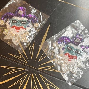 May include: Two jester face patches crafted with beads and sequins. Each patch features a purple hat, a white face with a red mouth, and a teal eye mask. The jester wears a white ruffled collar made of sequins. Both patches are sealed in clear plastic.