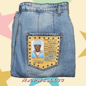 May include: Blue denim jeans featuring a back pocket with a tan rectangular patch printed to resemble a travel license. The patch includes a portrait of a person in a blue hat, the text Tyler Baudelaire, and a border of black stars.
