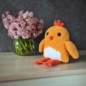 May include: A crocheted chick toy with orange and white yarn. The chick has a small orange comb and black eyes. The chick is sitting on a light brown surface next to a vase of pink hyacinths.