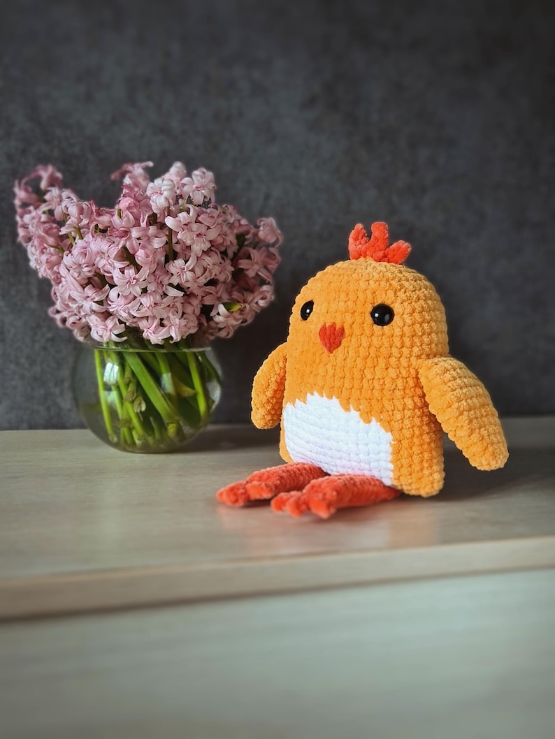 May include: A crocheted chick toy with orange and white yarn. The chick has a small orange comb and black eyes. The chick is sitting on a light brown surface next to a vase of pink hyacinths.