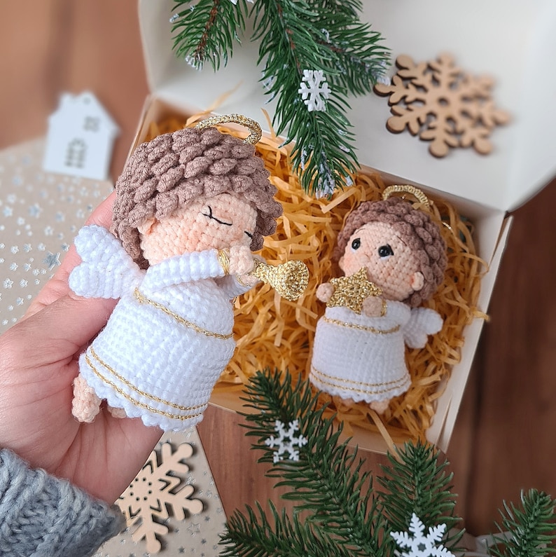 May include: Two crocheted angel ornaments in white with gold accents. The angels are holding a trumpet and a star. The ornaments are in a box with shredded paper.