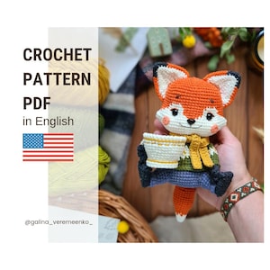Puede incluir: Patrón de crochet PDF en inglés para un adorable zorro amigurumi naranja que sostiene una taza. El zorro lleva un suéter verde y amarillo, jeans azules y una bufanda. El patrón está disponible para descargar.