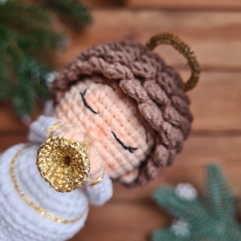 May include: A crocheted angel ornament with a gold halo and a golden trumpet. The angel has brown hair and closed eyes.