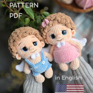 Könnte beinhalten: Zwei handgefertigte Engelspuppen aus Häkelarbeit mit braunem Haar, blauen Augen und kleinen weißen Flügeln. Eine Puppe trägt eine blaue Latzhose, die andere ein rosa Kleid. Der Text "CROCHET PATTERN PDF" und "in English" mit der amerikanischen Flagge ist enthalten.