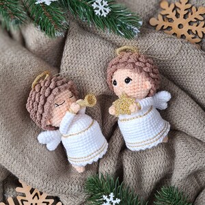May include: Two crocheted angel ornaments with gold accents. One angel is holding a trumpet and the other is holding a star. The angels are on a brown fabric background.