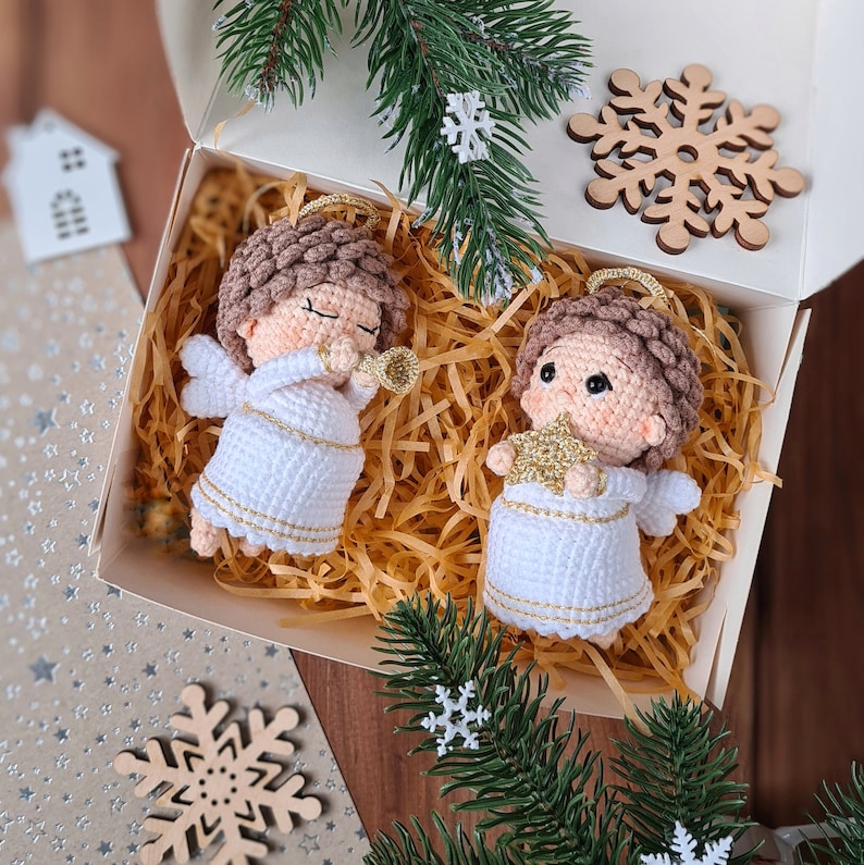 May include: Two white crocheted angel ornaments with gold trim and brown hair. The angels are holding a gold star and a trumpet. The ornaments are sitting in a box with shredded paper.