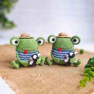May include: Two green crocheted frog plushies with straw hats and blue and white striped shirts. Each frog is holding a smaller crocheted fly.