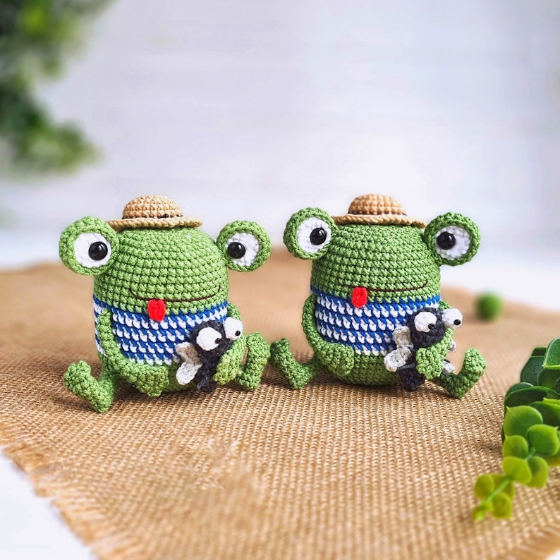 May include: Two green crocheted frog plushies with straw hats and blue and white striped shirts. Each frog is holding a smaller crocheted fly.