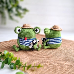 May include: Two crocheted green frog plushies with brown straw hats. The frog on the left is holding a smaller crocheted frog plushie. Both frogs are wearing blue and white striped shirts.