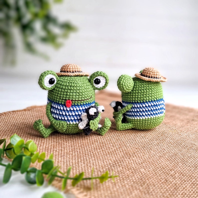 May include: Two crocheted green frog plushies with brown straw hats. The frog on the left is holding a smaller crocheted frog plushie. Both frogs are wearing blue and white striped shirts.