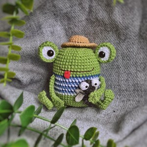 May include: A green crocheted frog wearing a brown hat and holding a small black and white crocheted bug. The frog has a red tongue sticking out.