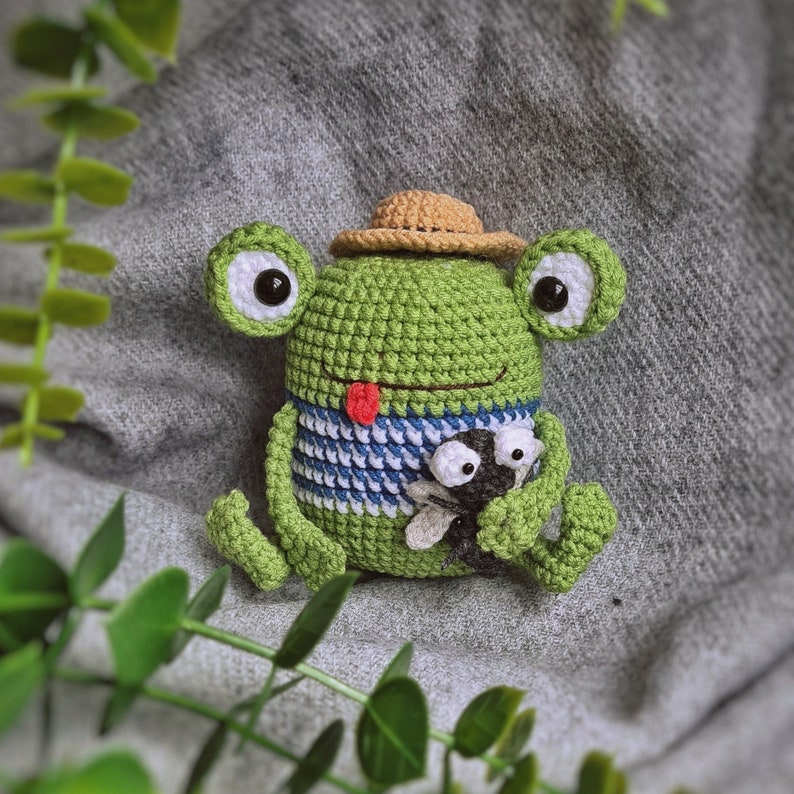 May include: A green crocheted frog wearing a brown hat and holding a small black and white crocheted bug. The frog has a red tongue sticking out.