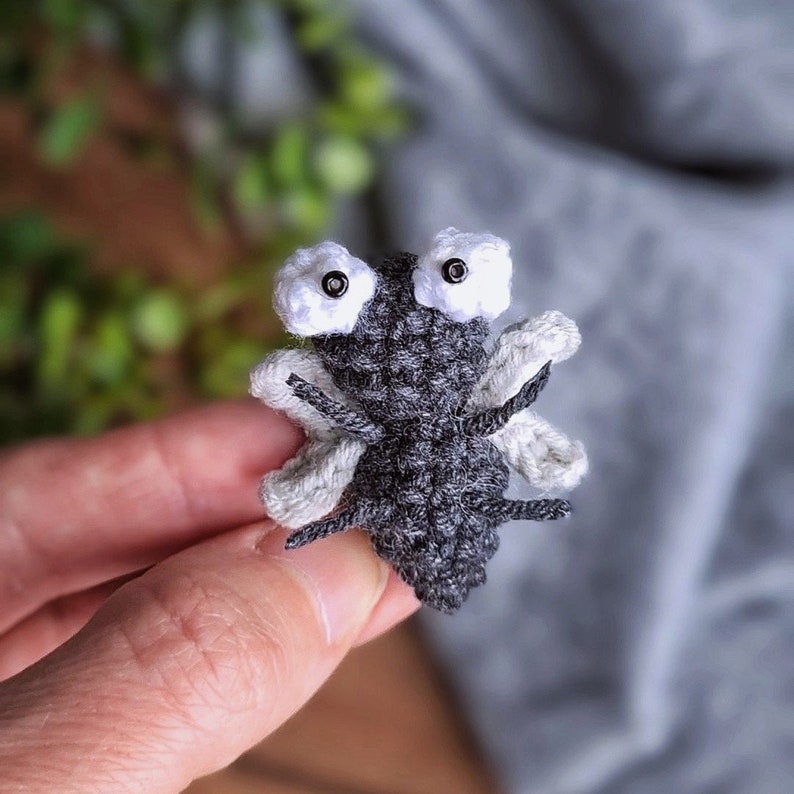 May include: A small crocheted fly with gray wings and white eyes. The fly is sitting on a hand.