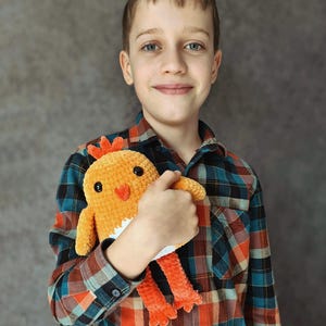 May include: A young person wearing a red and blue plaid shirt is holding a crocheted orange and white chick toy.