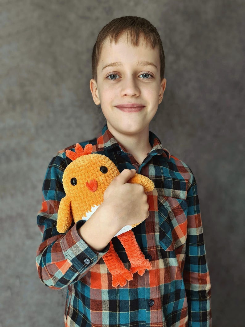 May include: A young person wearing a red and blue plaid shirt is holding a crocheted orange and white chick toy.