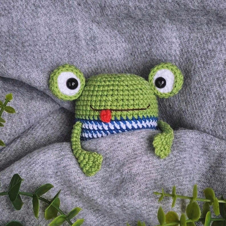 May include: A green crocheted frog with black eyes and a red tongue, wearing a blue and white striped scarf, is lying on a gray blanket.