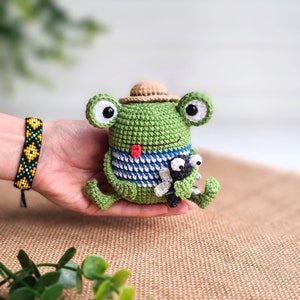 May include: A green crocheted frog wearing a brown hat holds a small black crocheted bug. The frog has a red mouth and blue and white striped shirt.