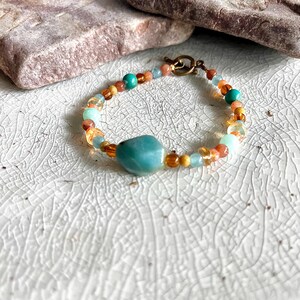 Peut inclure: Un bracelet de perles colorées avec une grosse pierre turquoise. Le bracelet est composé de perles de différentes couleurs : turquoise, orange et or. Il est doté d'un fermoir à crochet et œillet doré. Longueur approximative : 18 cm.