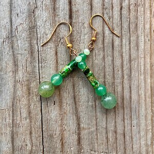 Op de afbeelding: Een paar handgemaakte oorbellen met goudkleurige oorhaken. Elke oorbel heeft een combinatie van groene en goudkleurige kralen, waaronder ronde en cilindrische vormen. De oorbellen worden gepresenteerd op een houten oppervlak.
