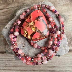 May include: A pink and brown gemstone pendant with gold flecks hangs from a multi-strand necklace of pink, red, and purple beads.