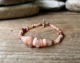 May include: A pink and brown gemstone bracelet with a copper clasp. The bracelet features a variety of different sized and shaped gemstones.