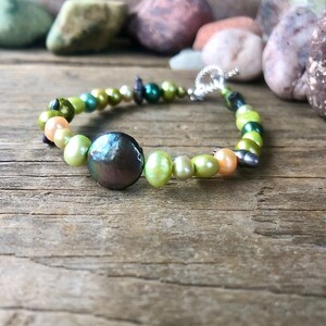 Peut inclure: Un bracelet en perles vertes, brunes et crème avec une grosse perle noire irisée au centre. Le bracelet est fait de petites perles rondes et a un fermoir argenté.
