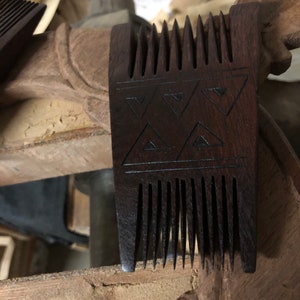 May include: A dark brown wooden comb with a handle and teeth. The handle features an engraved triangular pattern. The comb is likely handmade and appears to be a grooming tool.