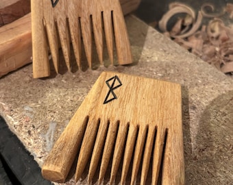 May include: Two wooden combs with a light brown finish and a black geometric symbol. The combs have a wide, rectangular top with evenly spaced teeth. The combs are handmade and appear to be crafted from a light-colored wood.