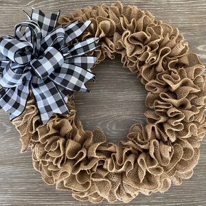 May include: A brown burlap wreath with a black and white checkered bow. The wreath is made of burlap fabric that has been gathered and ruffled to create a full and fluffy look. The bow is tied at the top of the wreath and is made of a black and white checkered ribbon.
