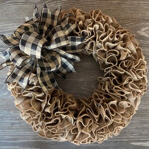 May include: A brown burlap wreath with a black and white checkered bow. The wreath is made of burlap and has a ruffled texture. The bow is tied in a knot and has a long tail.