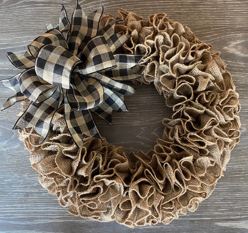 May include: A brown burlap wreath with a black and white checkered bow. The wreath is made of burlap fabric and has a ruffled texture. The bow is tied at the top of the wreath and has a large, flowing design.