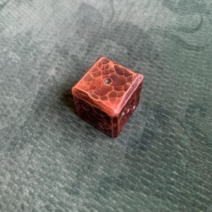 May include: A handcrafted copper die with a textured surface and visible markings. The die is a rich, reddish-brown colour and appears to be made of solid metal. The die is a cube shape.