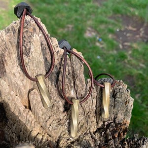 May include: Three brass pendant necklaces with brown leather cords are displayed on a weathered wooden surface. Each pendant has a twisted, rectangular design. The necklaces are set against a blurred green background.